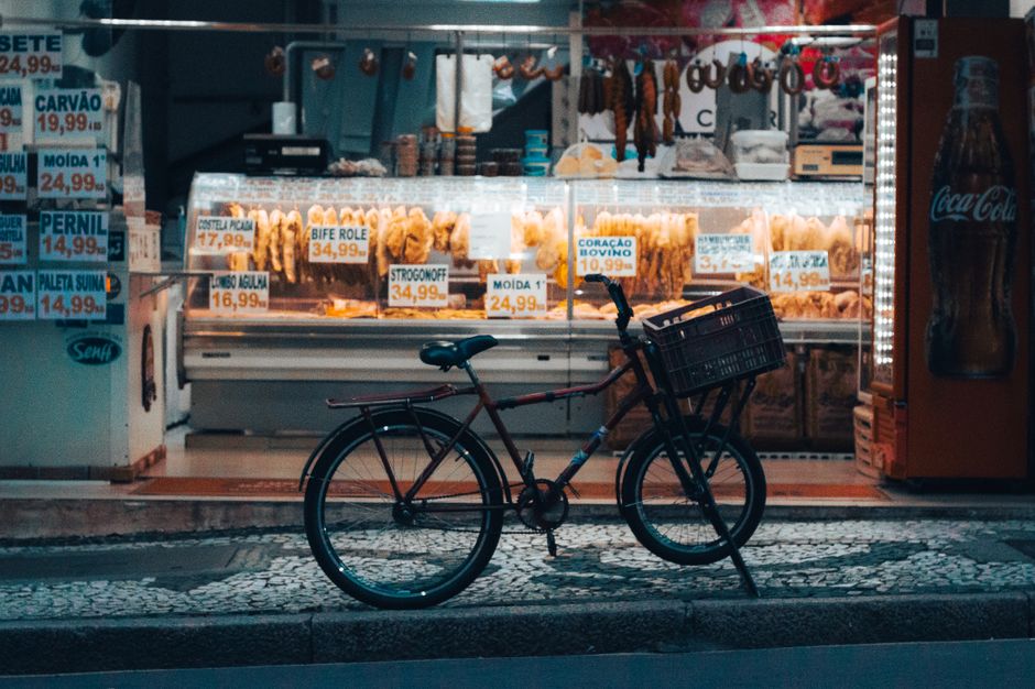Uma bicicleta estacionada do lado de fora de uma loja iluminada em Curitiba à noite, exibindo vários produtos do mercado